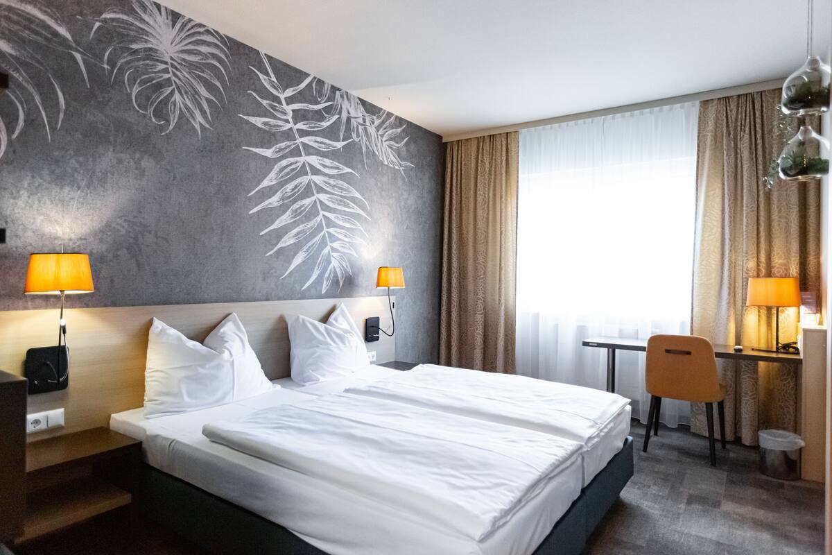 A hotel room with a large bed, two bedside lamps, a desk with a chair, and a window with curtains.