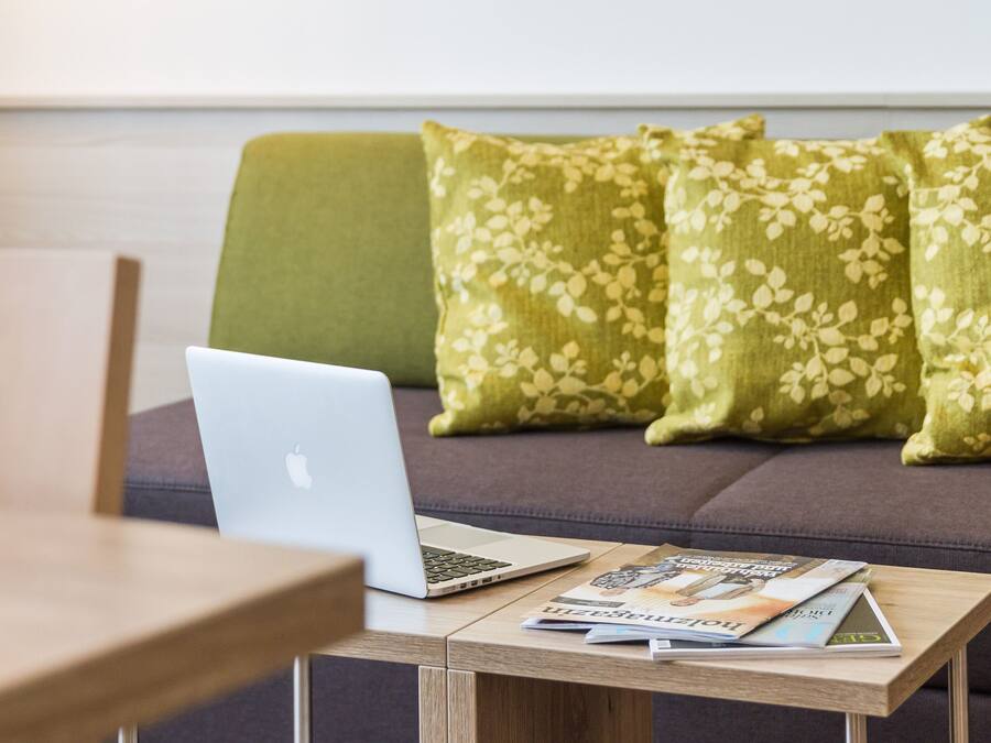 Una computadora portátil sobre una mesa de madera con revistas, un sofá gris con cojines estampados y una silla de madera.
