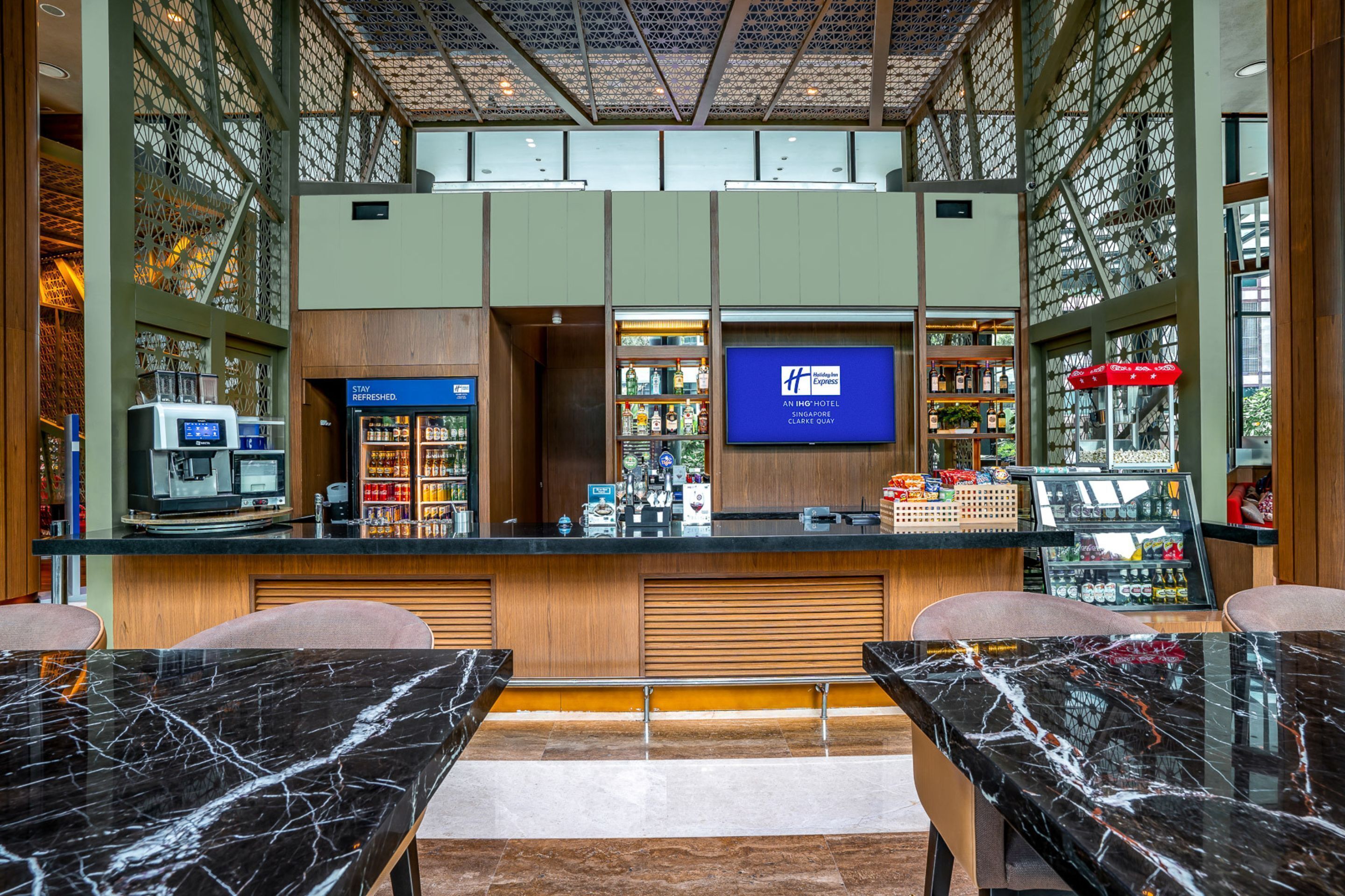 A hotel lobby with a reception desk, a minibar, seating area, and a television screen displaying the hotel name.