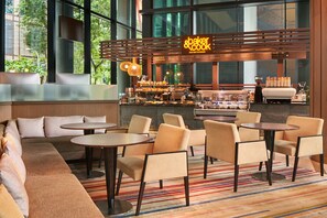 Un café moderno con zona de estar que incluye sofás y mesas redondas, una barra de café a la vista y grandes ventanales con vistas al exterior.