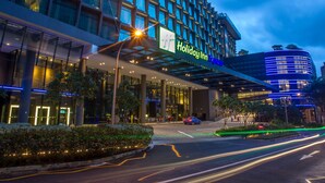 Edificio Holiday Inn Express con fachada de vidrio moderna, acceso cubierto y zona ajardinada con árboles y arbustos.