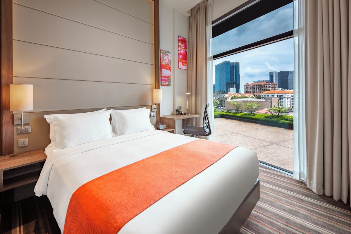 Habitación de hotel con una cama grande, un escritorio, una silla y vistas a la ciudad a través de un ventanal amplio.