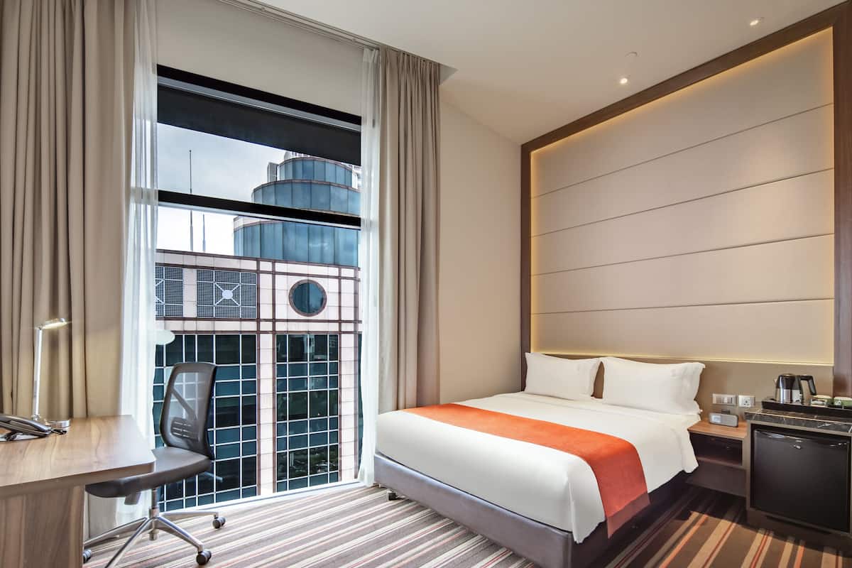 Habitación de hotel con una cama grande, un escritorio, una silla y vista a un edificio a través de una ventana.