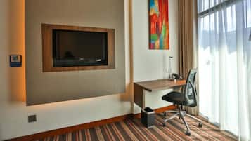 A hotel room with a flat-screen TV mounted on the wall, a desk with a chair, and a window with curtains.
