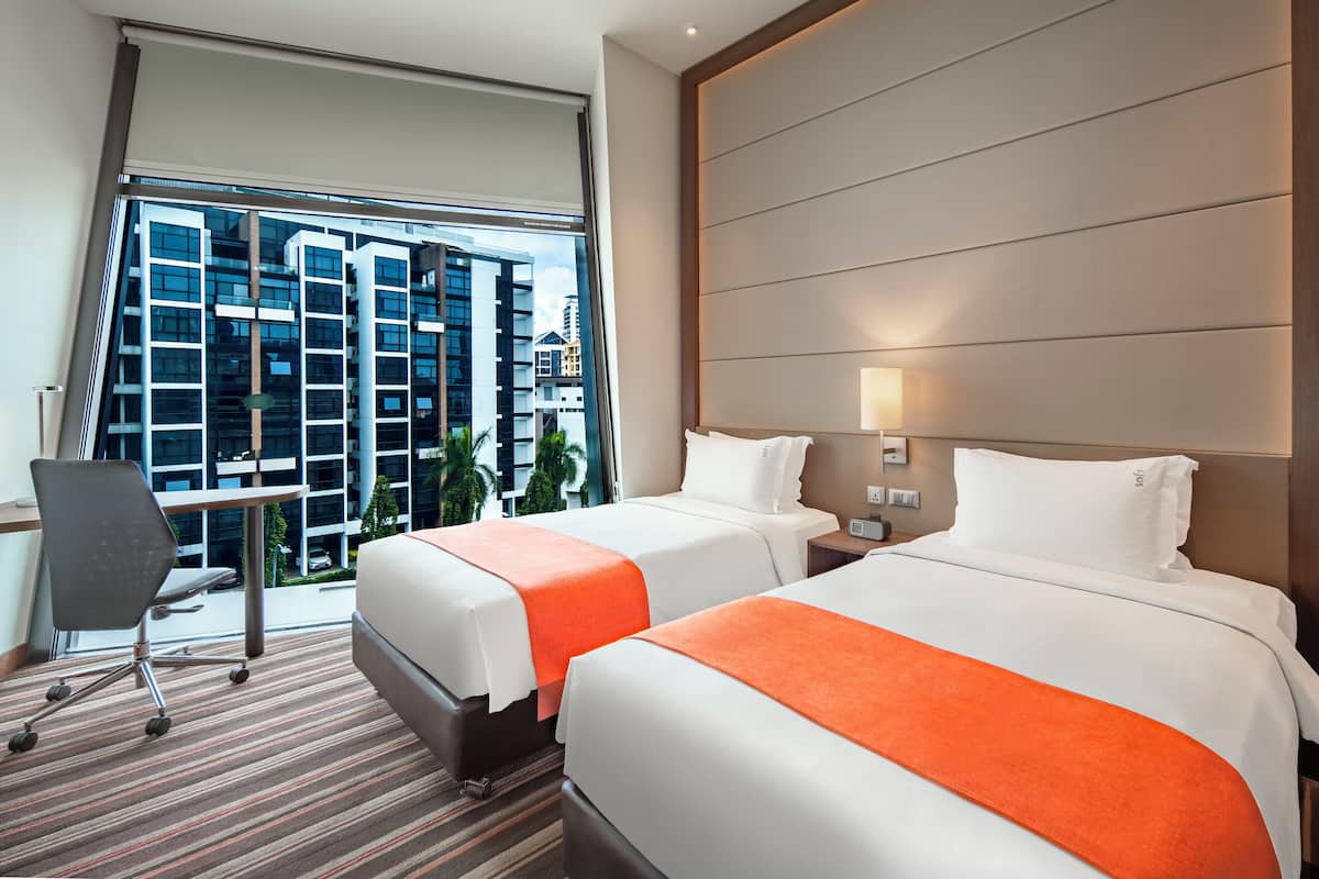 Habitación de hotel con dos camas, un escritorio y una silla. La habitación tiene un ventanal con vistas a un edificio alto.