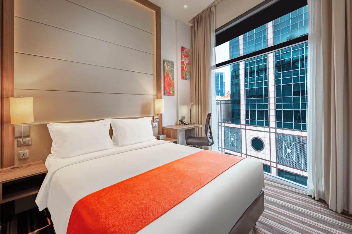 Una habitación de hotel con una cama grande, un escritorio y vistas de la ciudad a través de amplias ventanas.