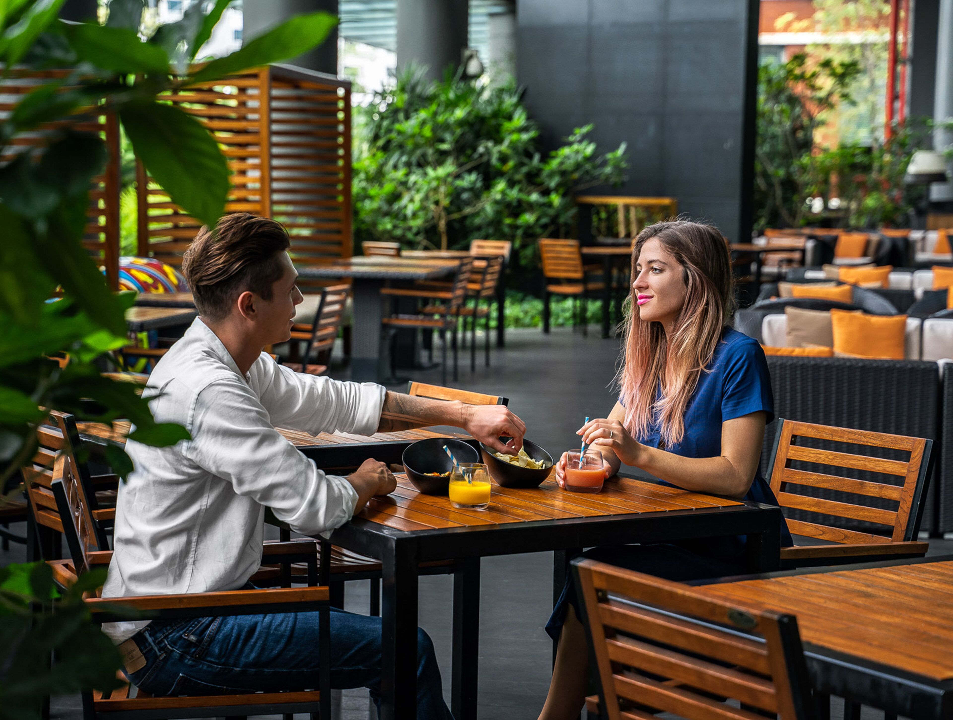 Una pareja cenando en un café al aire libre con mobiliario de madera y follaje de fondo.