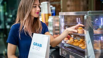 A person holding a white paper bag with 'GRAB & GO' text, standing in front of a bakery display case with pastries.