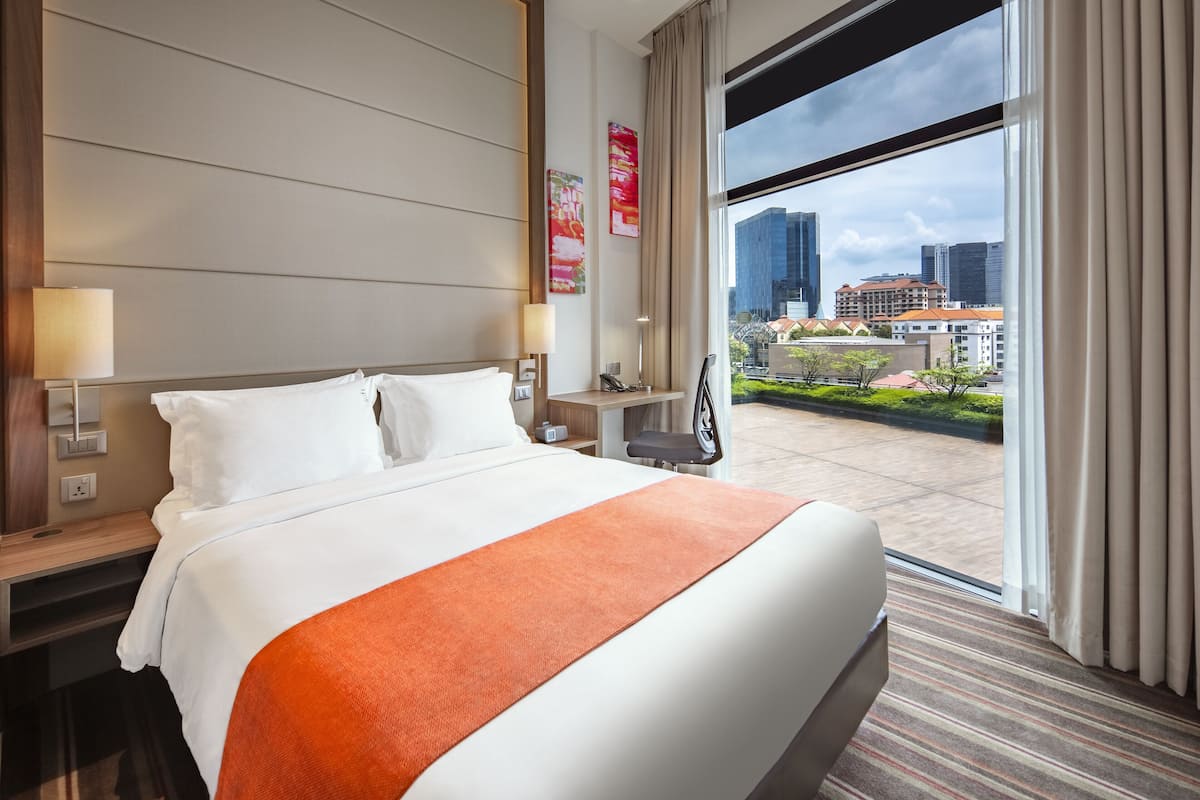 Habitación de hotel con una cama grande, un escritorio, una silla y vistas a la ciudad a través de un ventanal amplio.