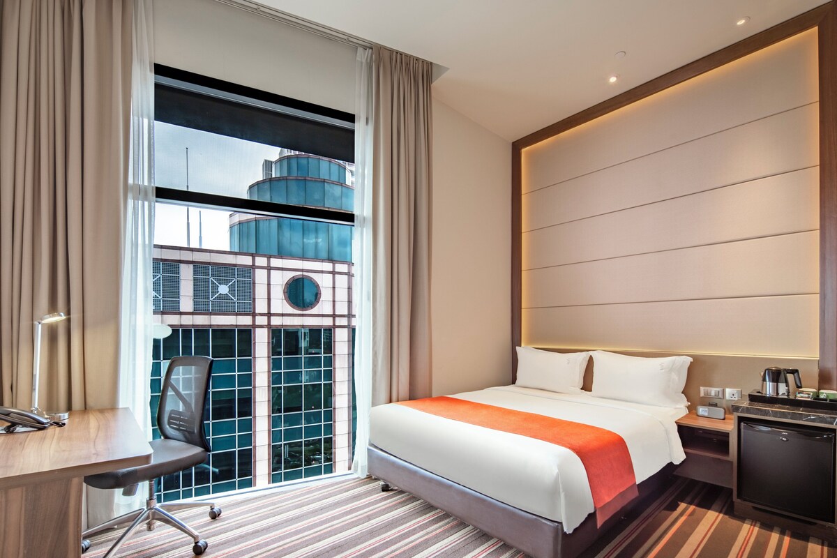 Habitación de hotel con una cama grande, un escritorio, una silla y vista a un edificio a través de una ventana.