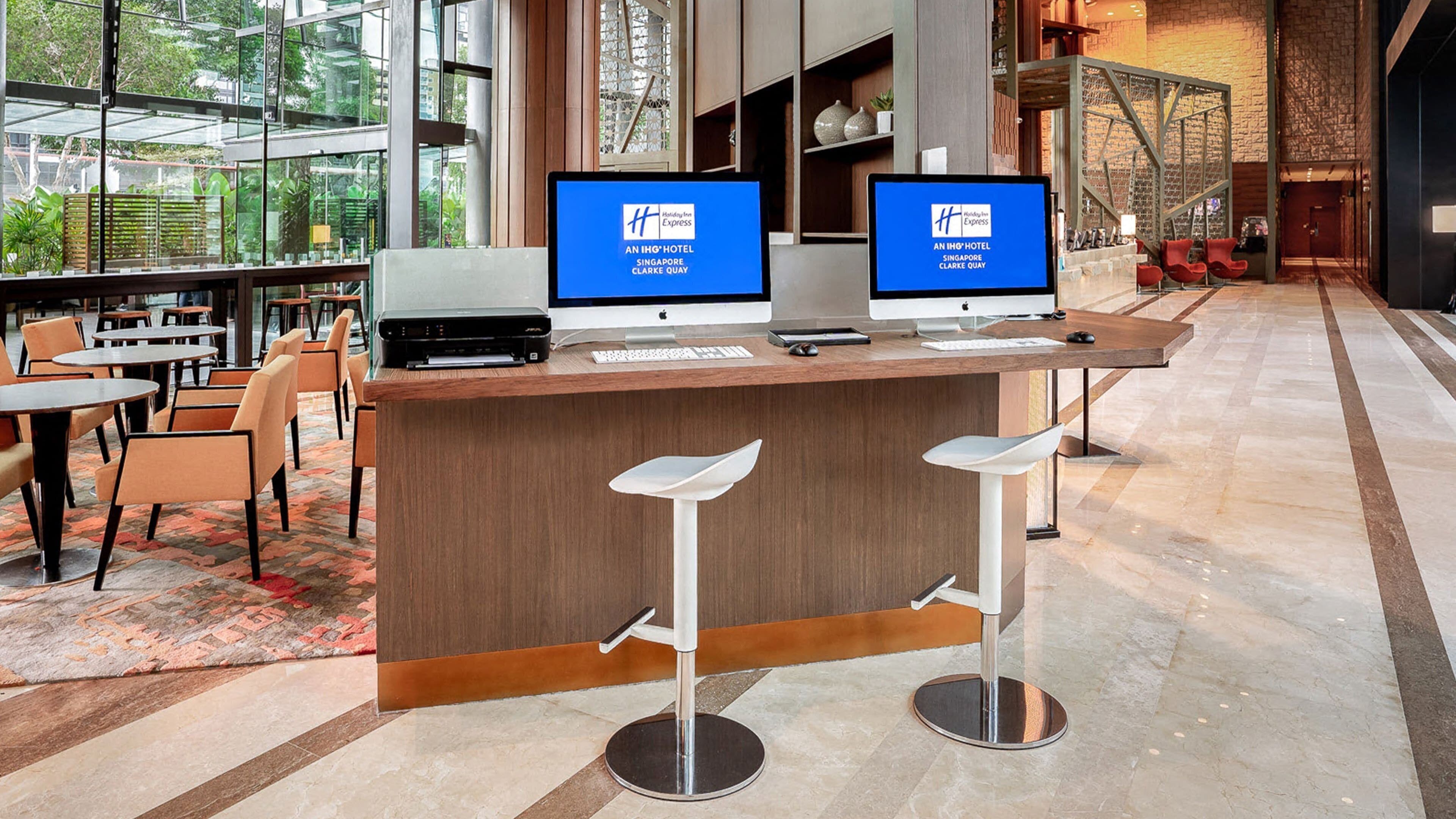 A hotel reception area with two monitors displaying the hotel’s logo, a wooden reception desk, and modern bar stools.