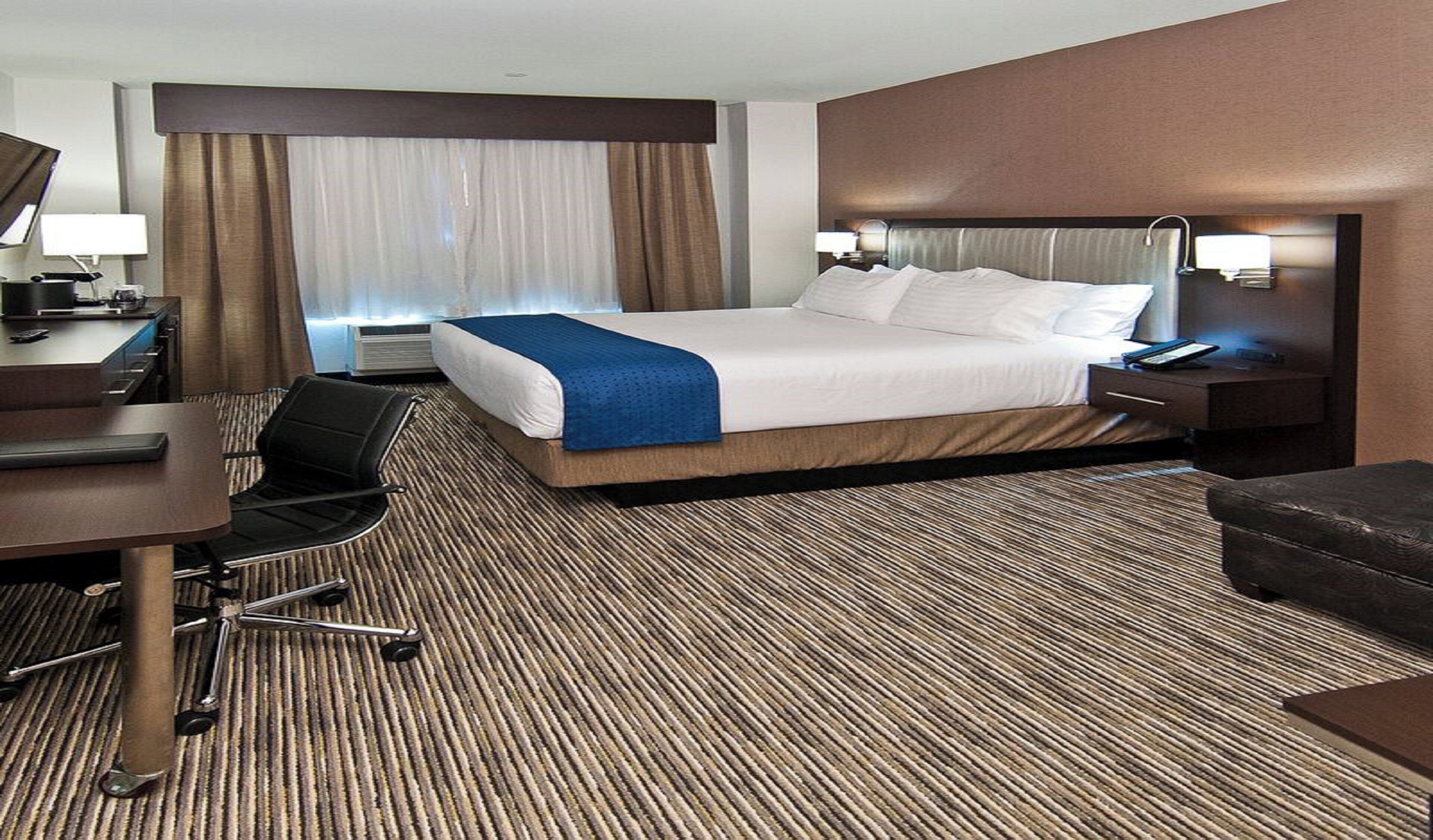 Habitación de hotel con cama, escritorio, silla y un amplio ventanal con cortinas.