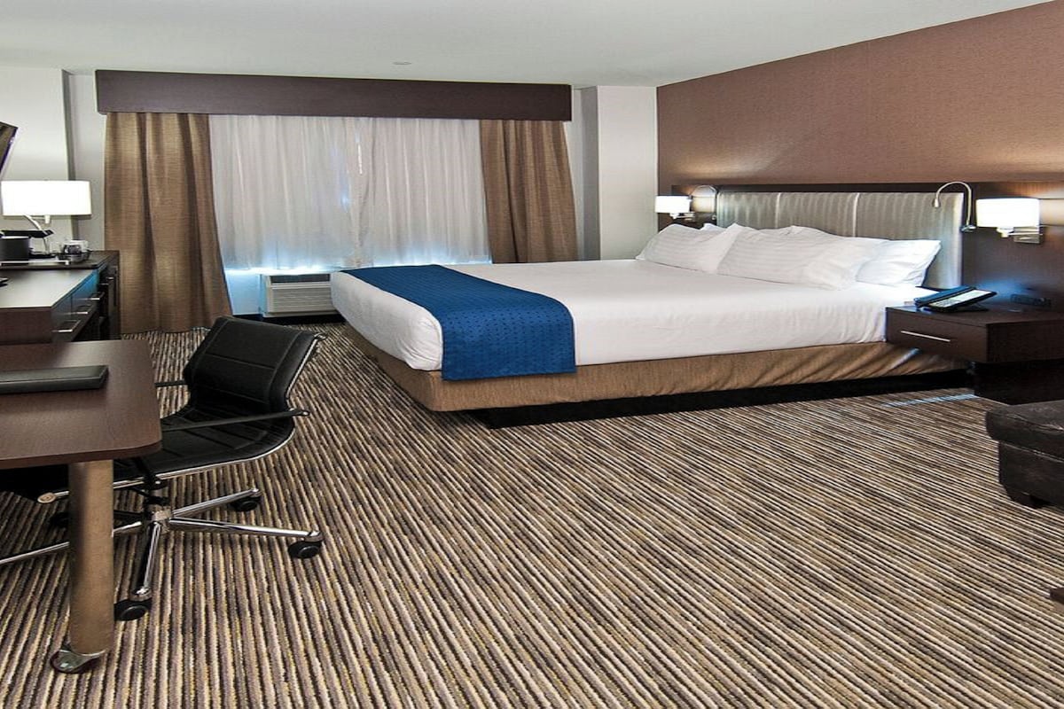 A hotel room with a bed, desk, chair, and a large window with curtains.