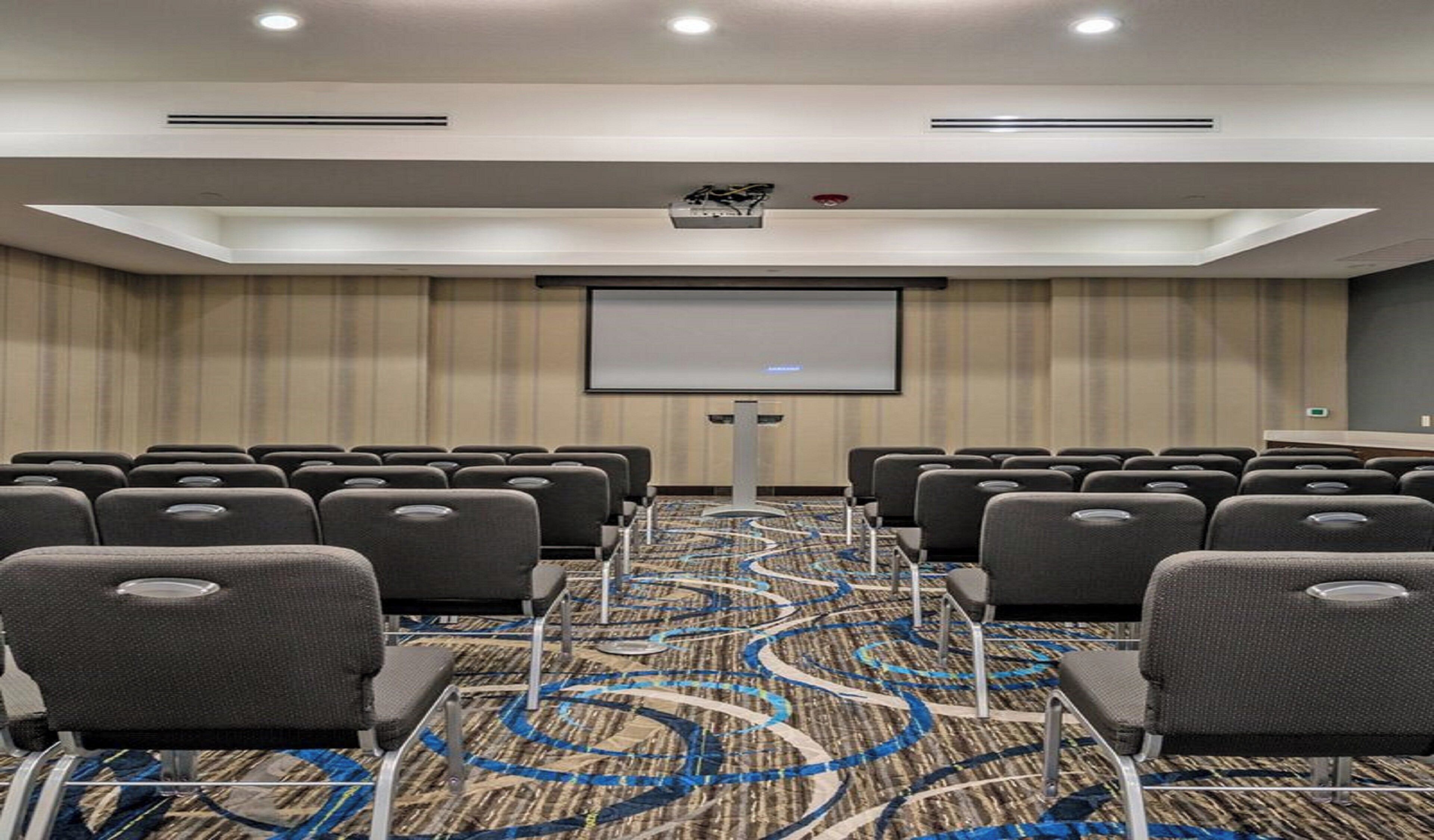 Un salón de conferencias con filas de sillas, una pantalla de proyector y piso alfombrado.