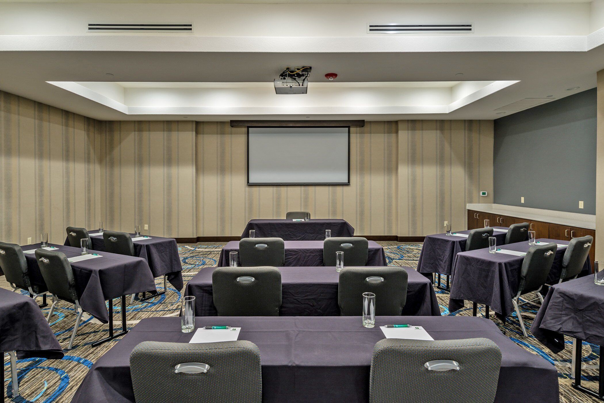 Una sala de conferencias con mesas y sillas, una pantalla de proyector y piso alfombrado.