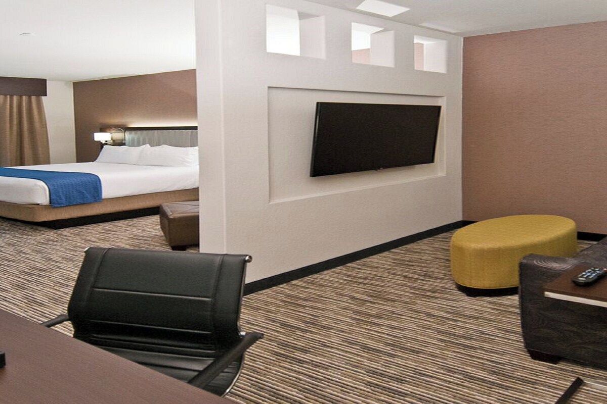 Een hotelkamer met een bed, een aan de muur bevestigde televisie, een bureau met stoel, en een zithoek met een geel pouf.