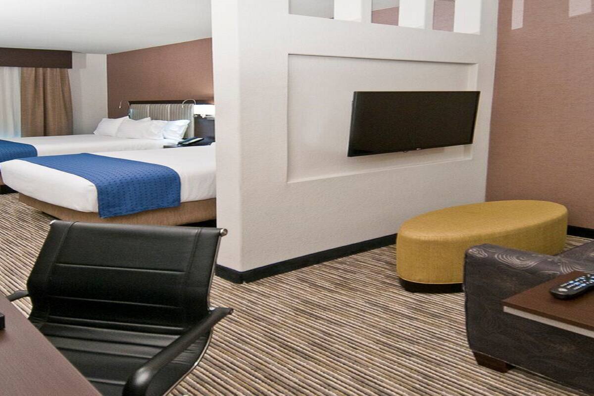 A hotel room with two beds, a TV mounted on the wall, a desk with a chair, and a small ottoman.