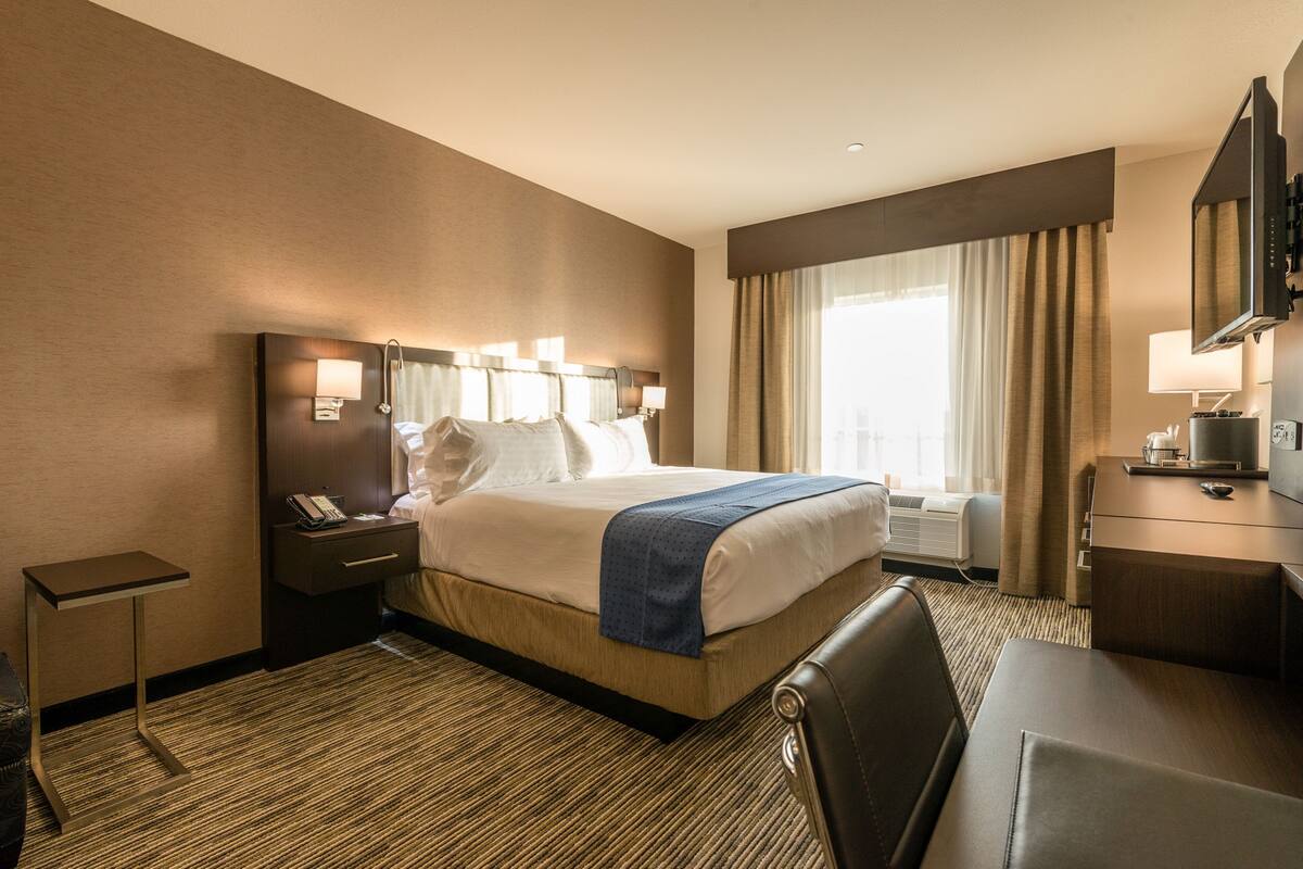 A hotel room with a large bed, bedside tables, a desk, a chair, a TV, and a window with curtains.