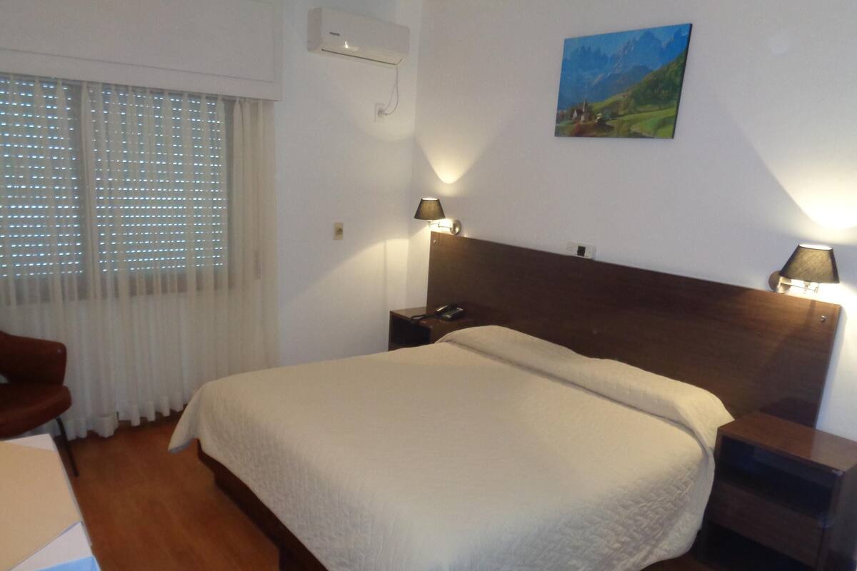 Hotellihuone, jossa on sänky, yöpöytävalaisimet, taulu seinällä ja ikkuna, jossa on sälekaihtimet.