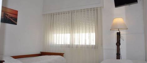 Quarto com duas camas de solteiro, janela com cortinas leves, luminária de cabeceira, televisão de tela plana e ar condicionado.
