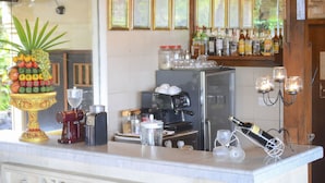 Una zona de bar con techo de madera, luces colgantes y una barra con decoración de piña. Hay carteles en la pared y varios botellas y vasos en las estanterías.