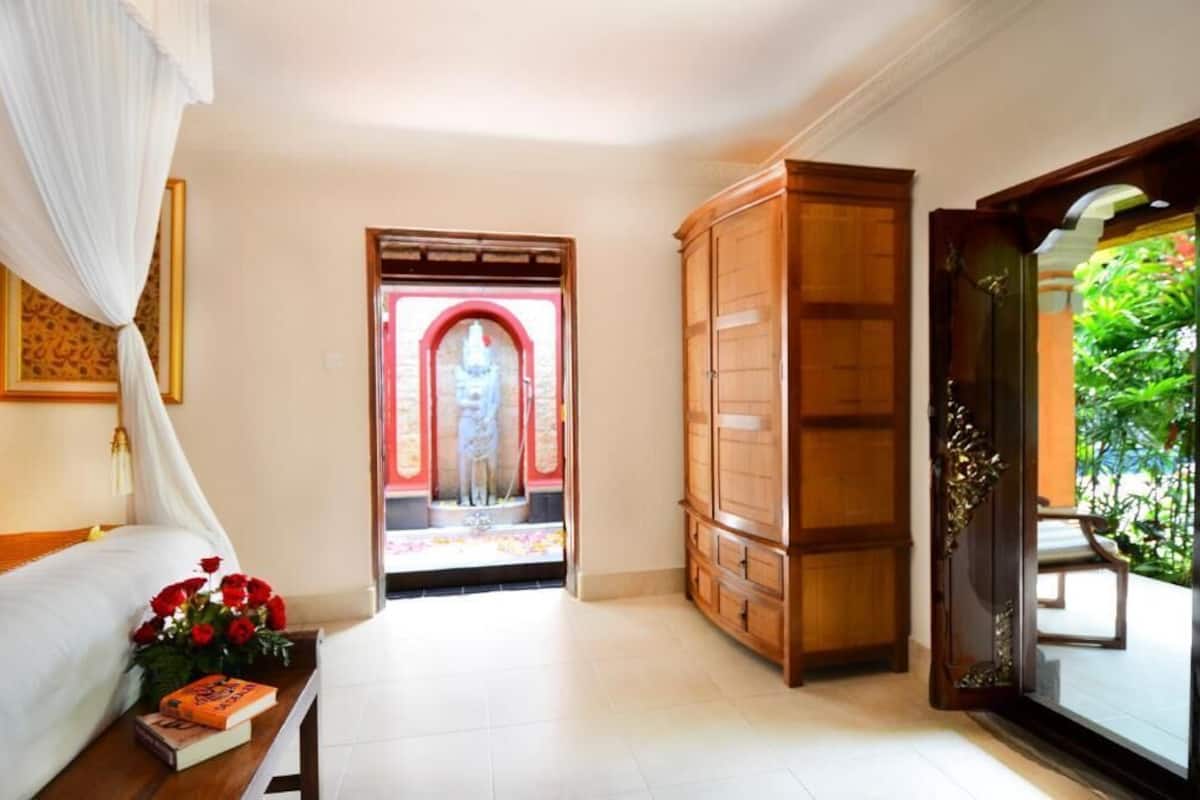 A hotel room with a bed, bedside table with books and flowers, a wooden wardrobe, and a view of a garden through an open door.