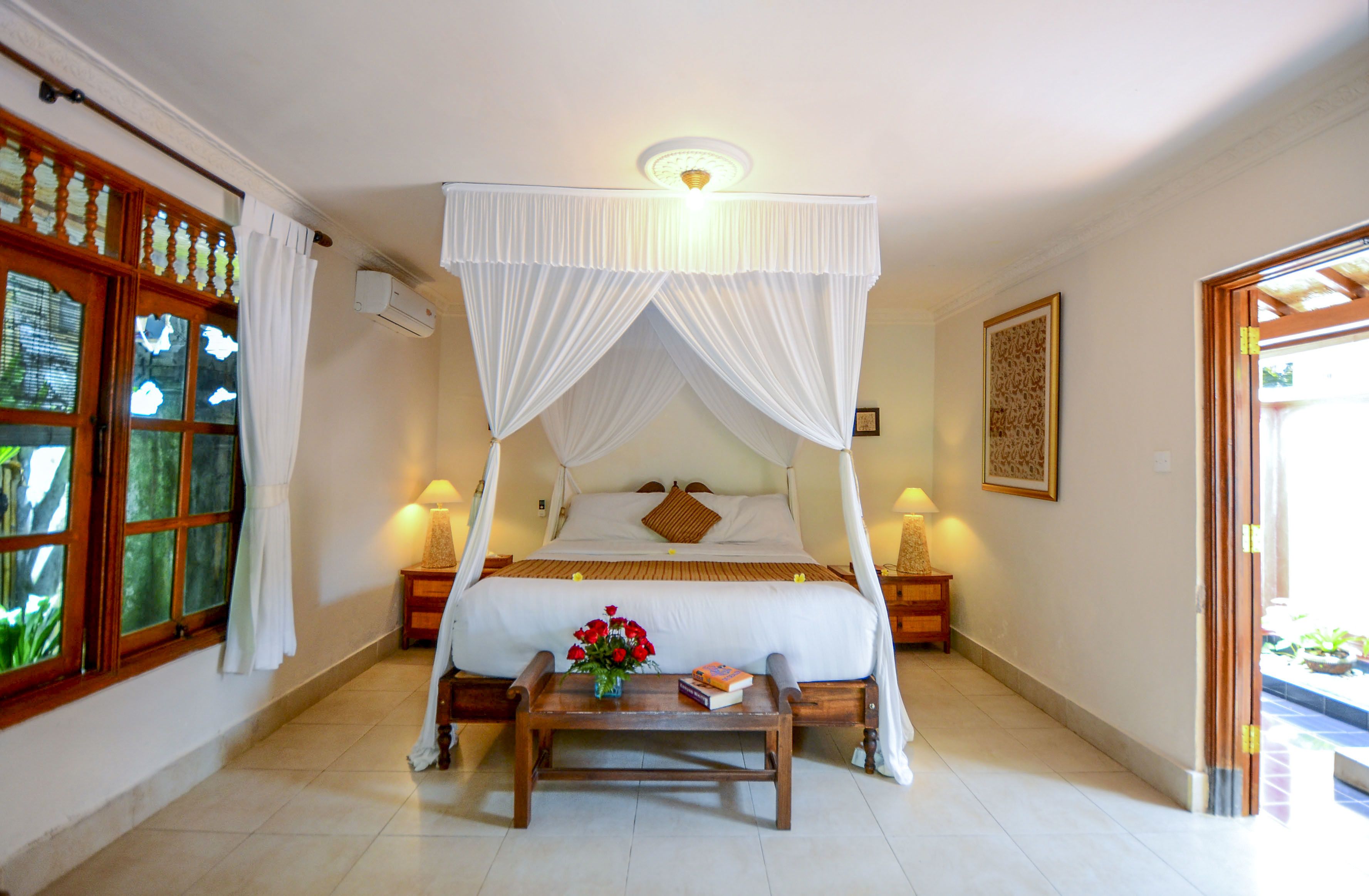 A four-poster bed with white drapes, a bedside table with a lamp, and a window with curtains.