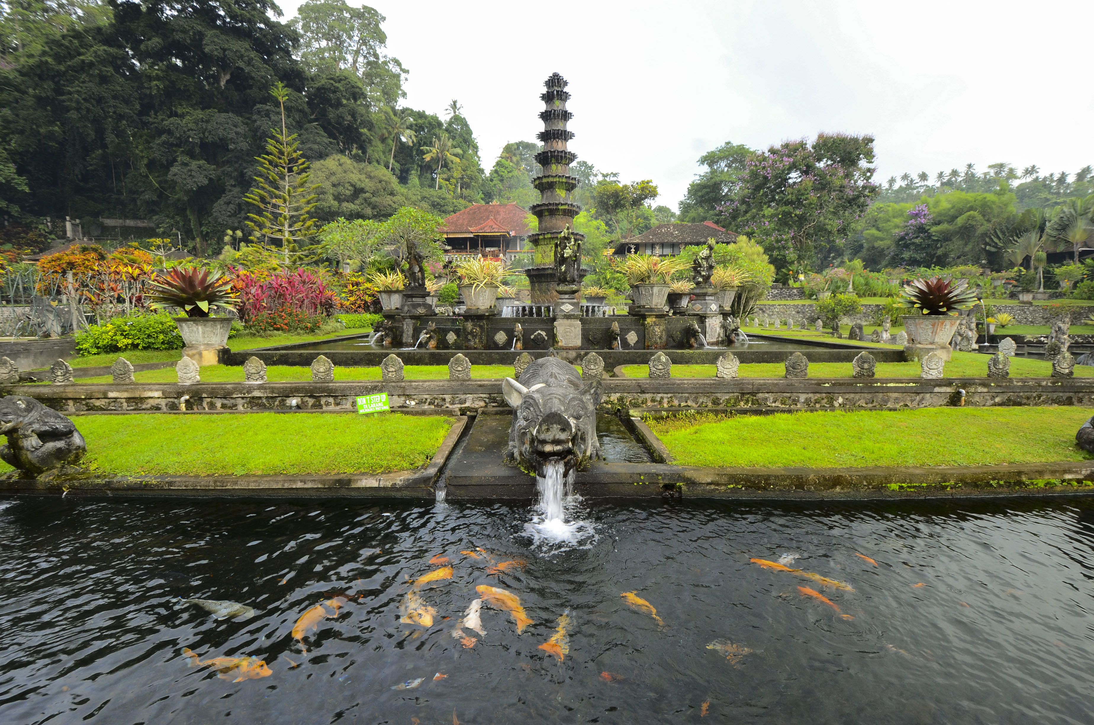 A traditional stone fountain with a tiered structure and a central water spout, surrounded by well-maintained greenery and a calm water surface with koi fish.