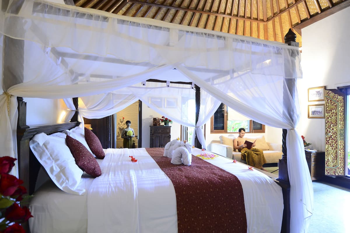 A bed with a canopy, a sofa, and a reading area in a room with a wooden ceiling and a view of another room.