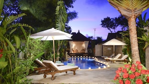 Zona de piscina con sillones de descanso y sombrillas, rodeada de plantas tropicales y un pabellón.