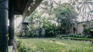 A lush garden with a stone structure, a thatched roof, and tropical plants. - Villa Puri Darma Agung (Ubud)