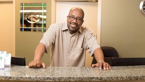 A man leaning on a reception desk in a hotel lobby with a framed sign on the wall and an Acer laptop on the desk. - Capital Suites Yellowknife (Yellowknife)