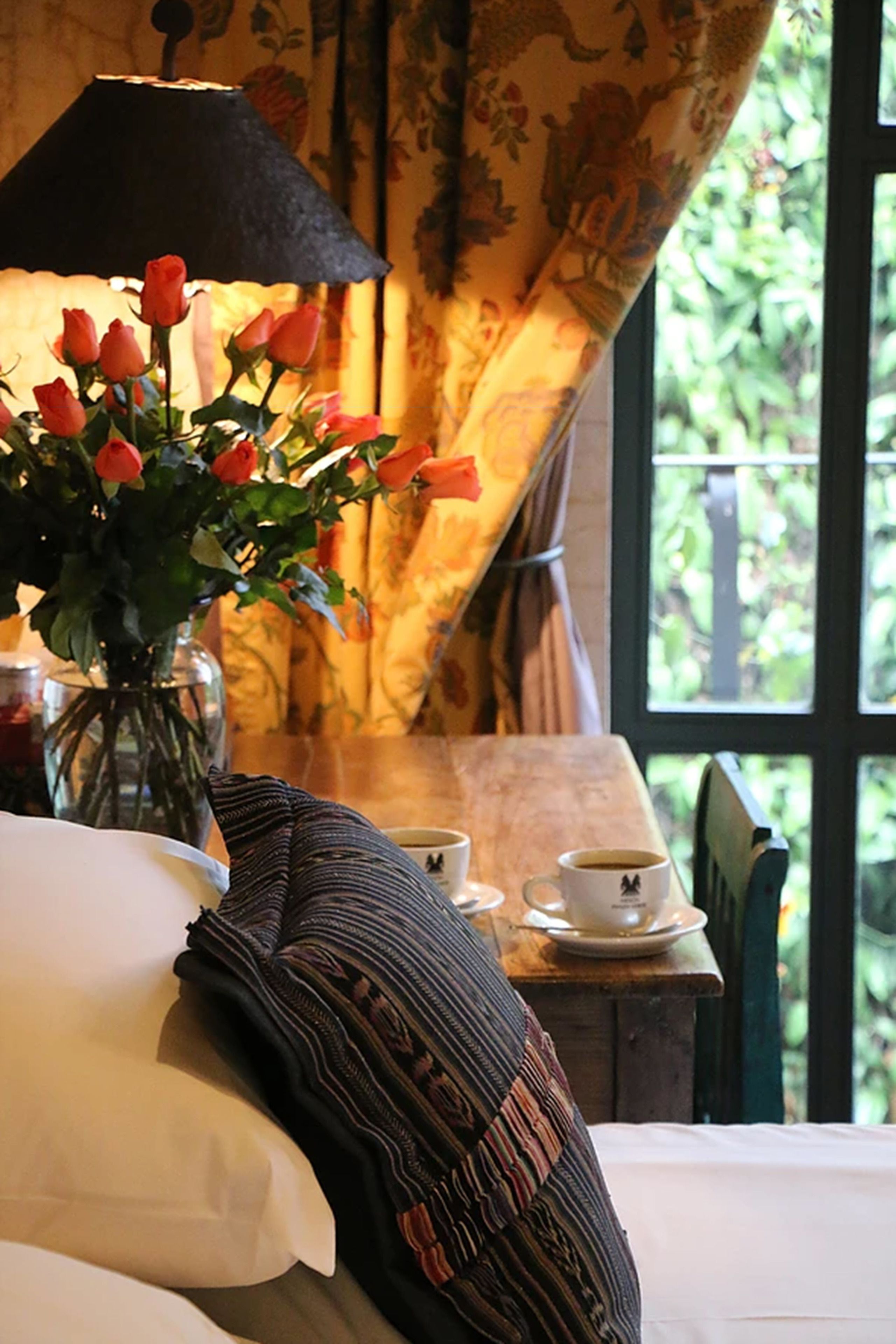 Una cama con un almohadón blanco y una colcha a rayas, un jarrón con rosas rojas, una mesa con una taza y platillo, y una ventana con vistas a la vegetación.