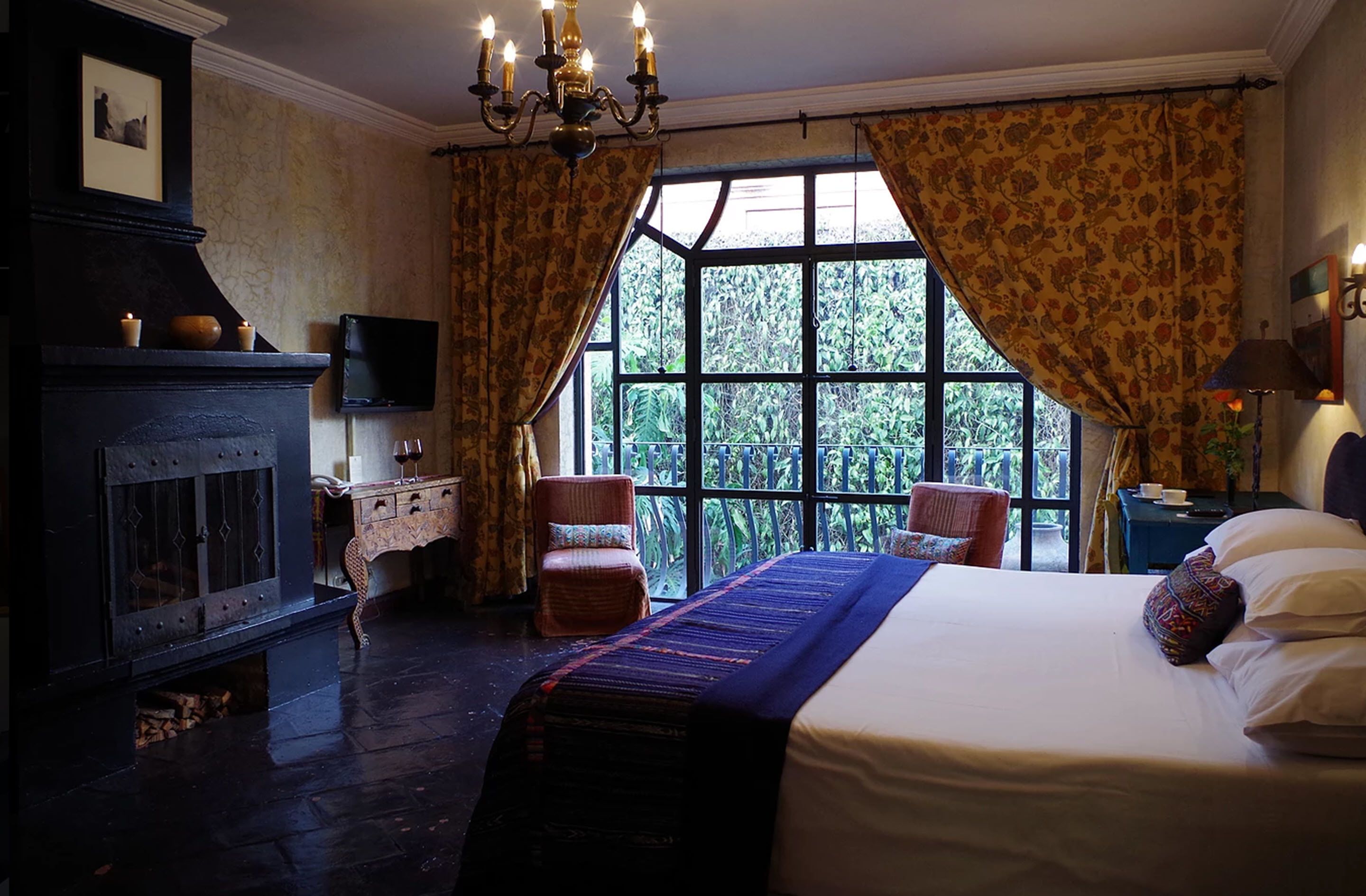 A bedroom with a large bed, a fireplace, a television, and a view of greenery through the window.