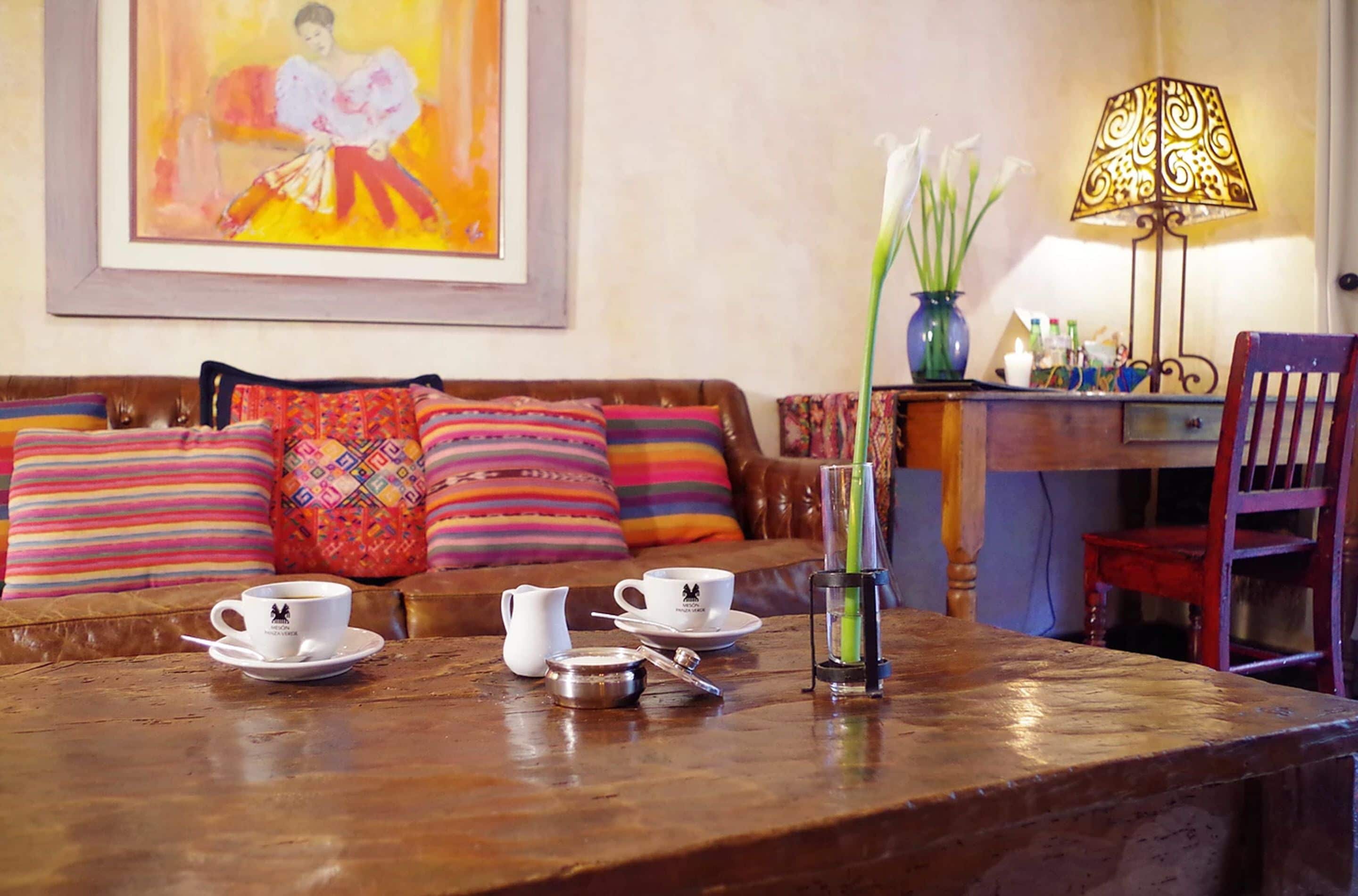 A cozy living area with a wooden coffee table, a sofa with colorful cushions, a framed painting on the wall, and a lamp.