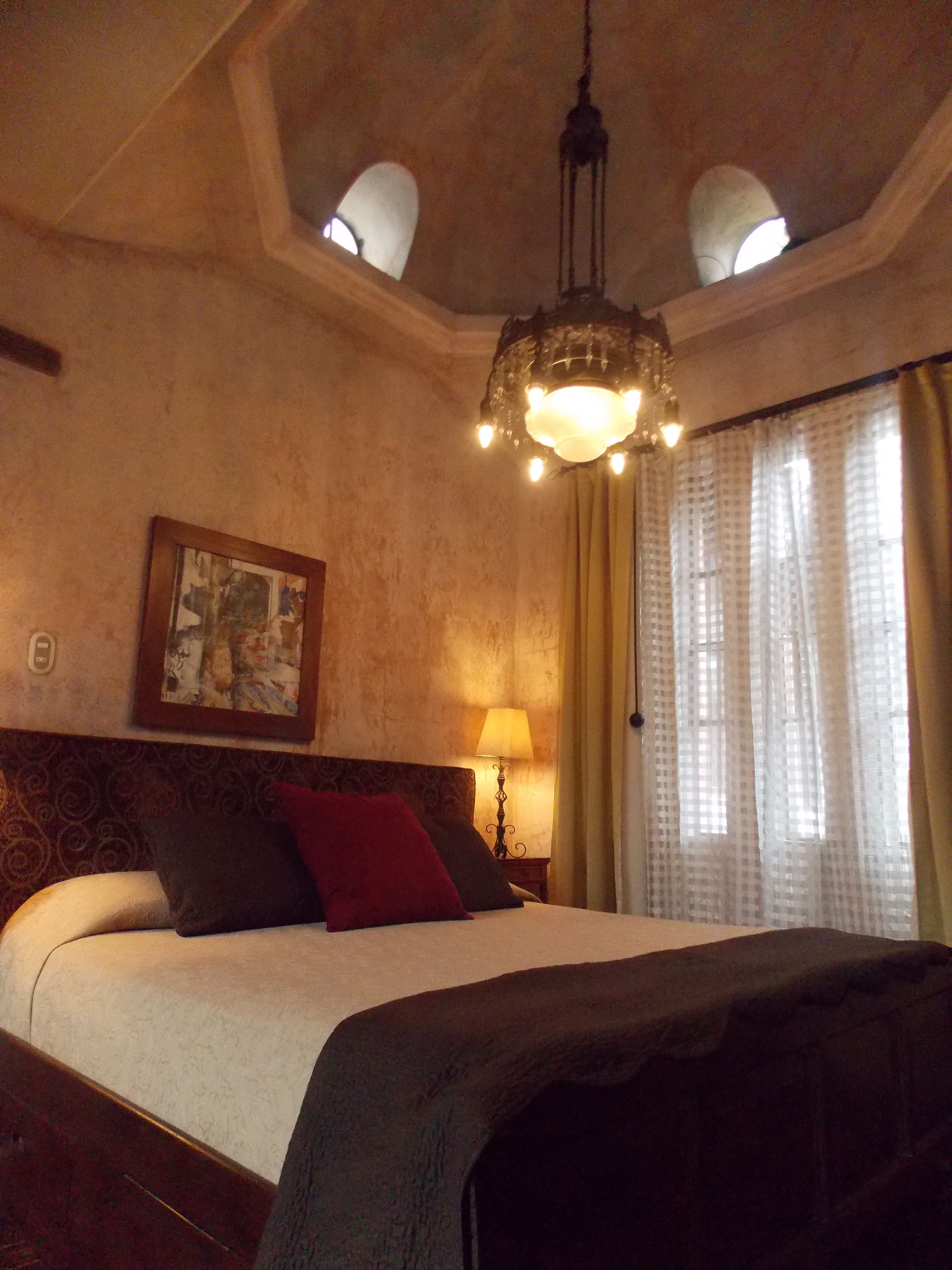 A hotel room with a large bed, a chandelier, a painting on the wall, and a window with curtains.