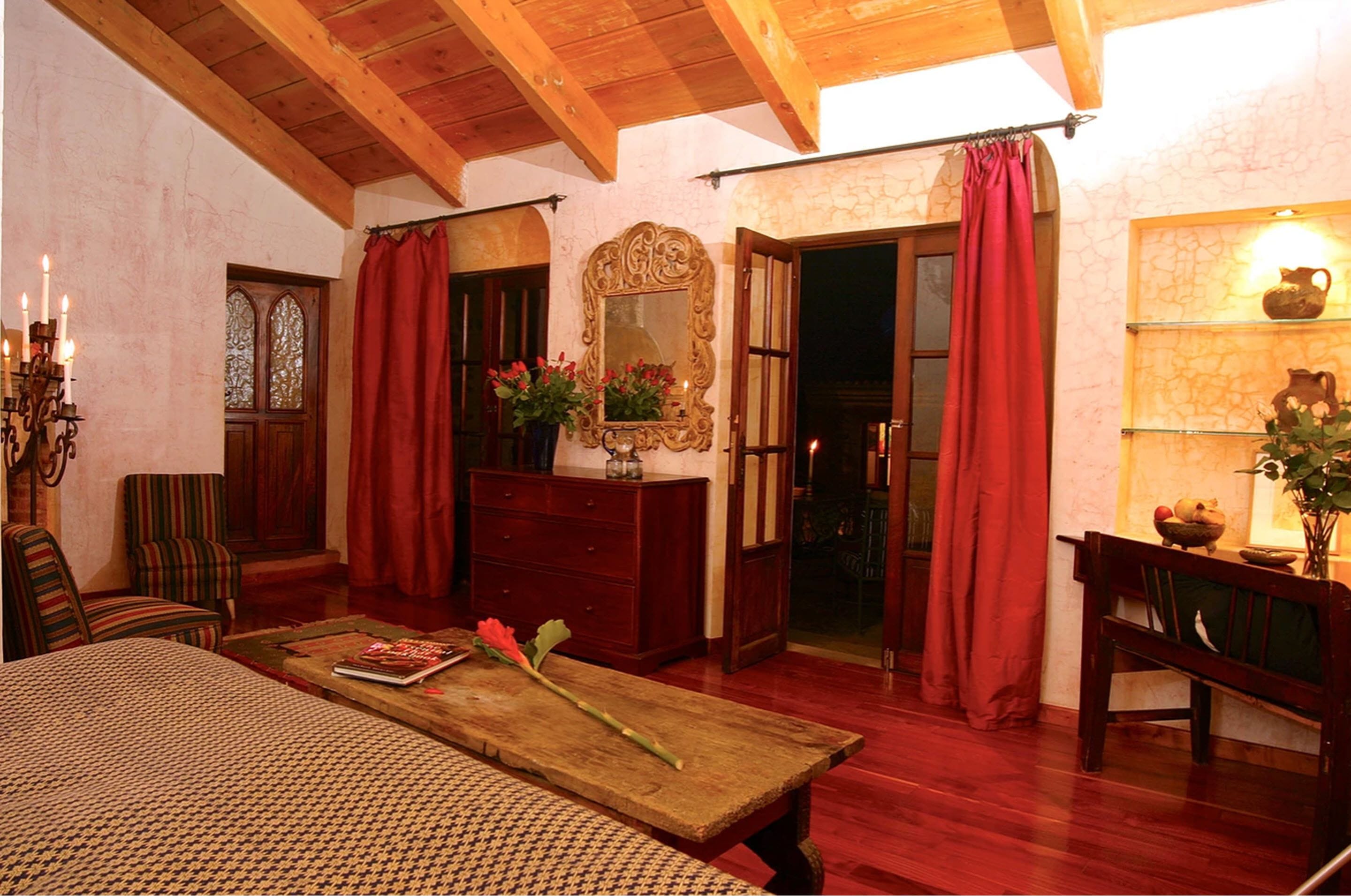 A cozy room with a wooden ceiling, a large bed, a wooden dresser, a mirror, and red curtains.