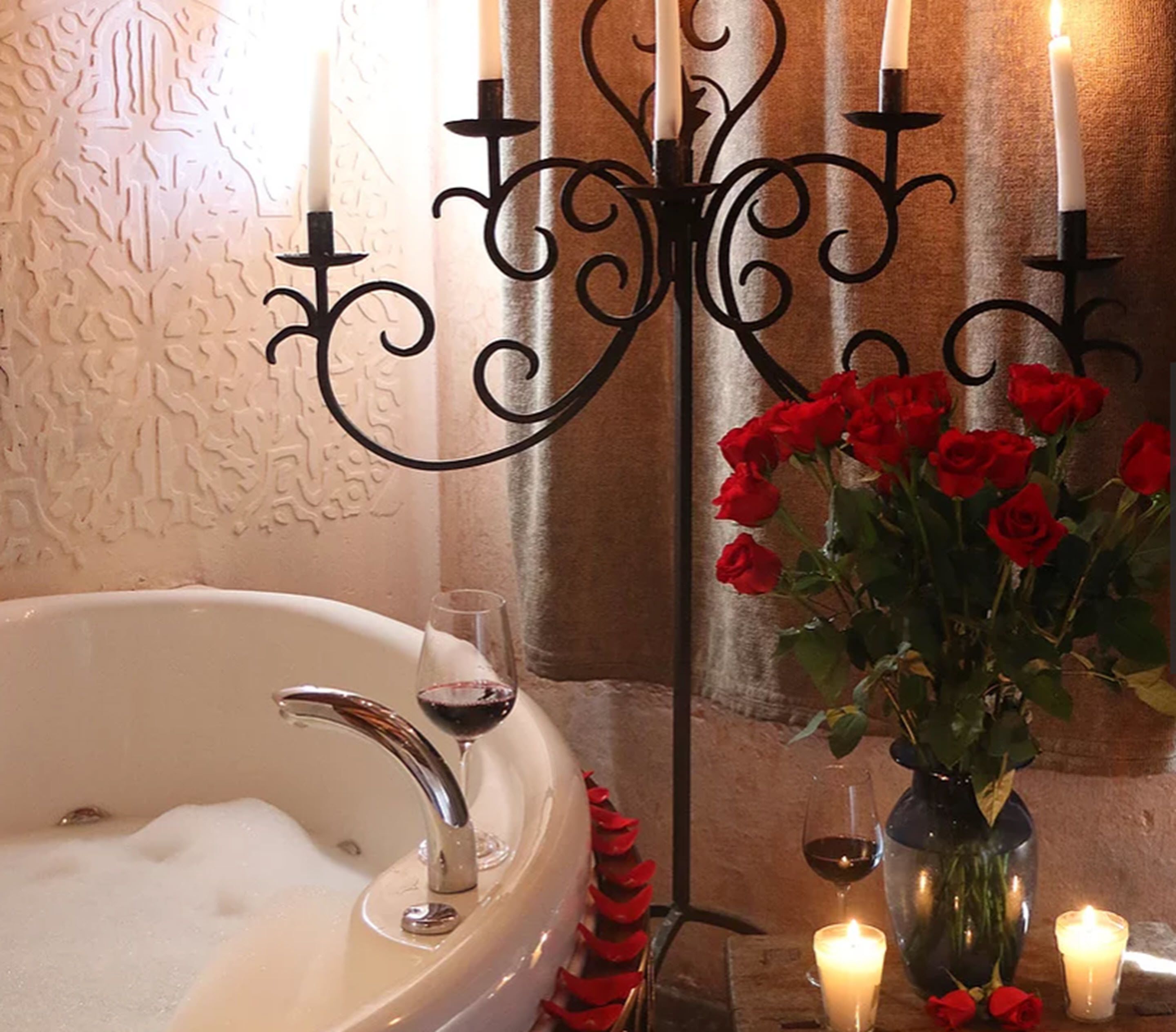 Ein Badezimmer mit einer Badewanne, einem Glas Rotwein, einer Vase mit roten Rosen und Kerzen.