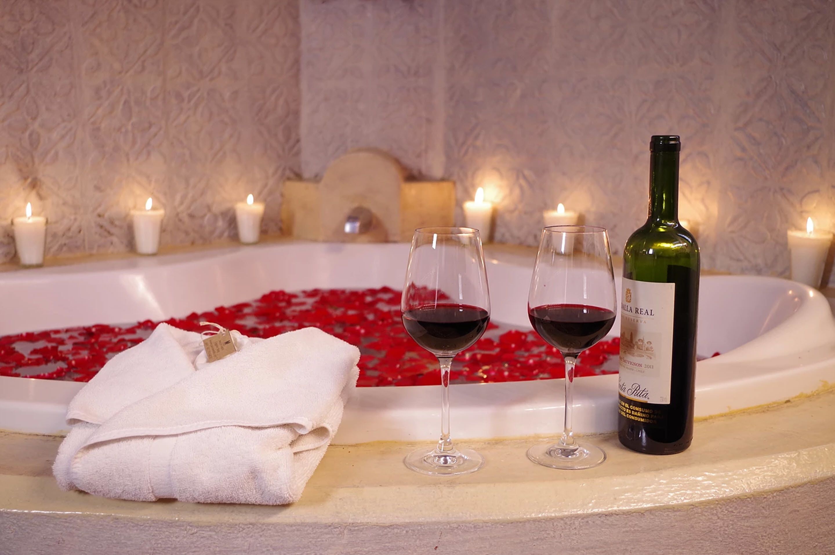 A bathtub filled with red rose petals, two wine glasses, a bottle of wine, and several lit candles.