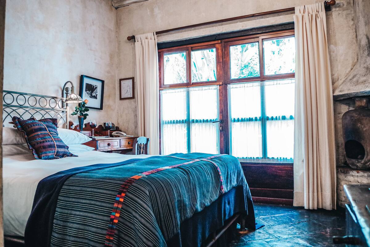 Une chambre à coucher avec un grand lit, une table de chevet, une lampe et une fenêtre avec des rideaux.