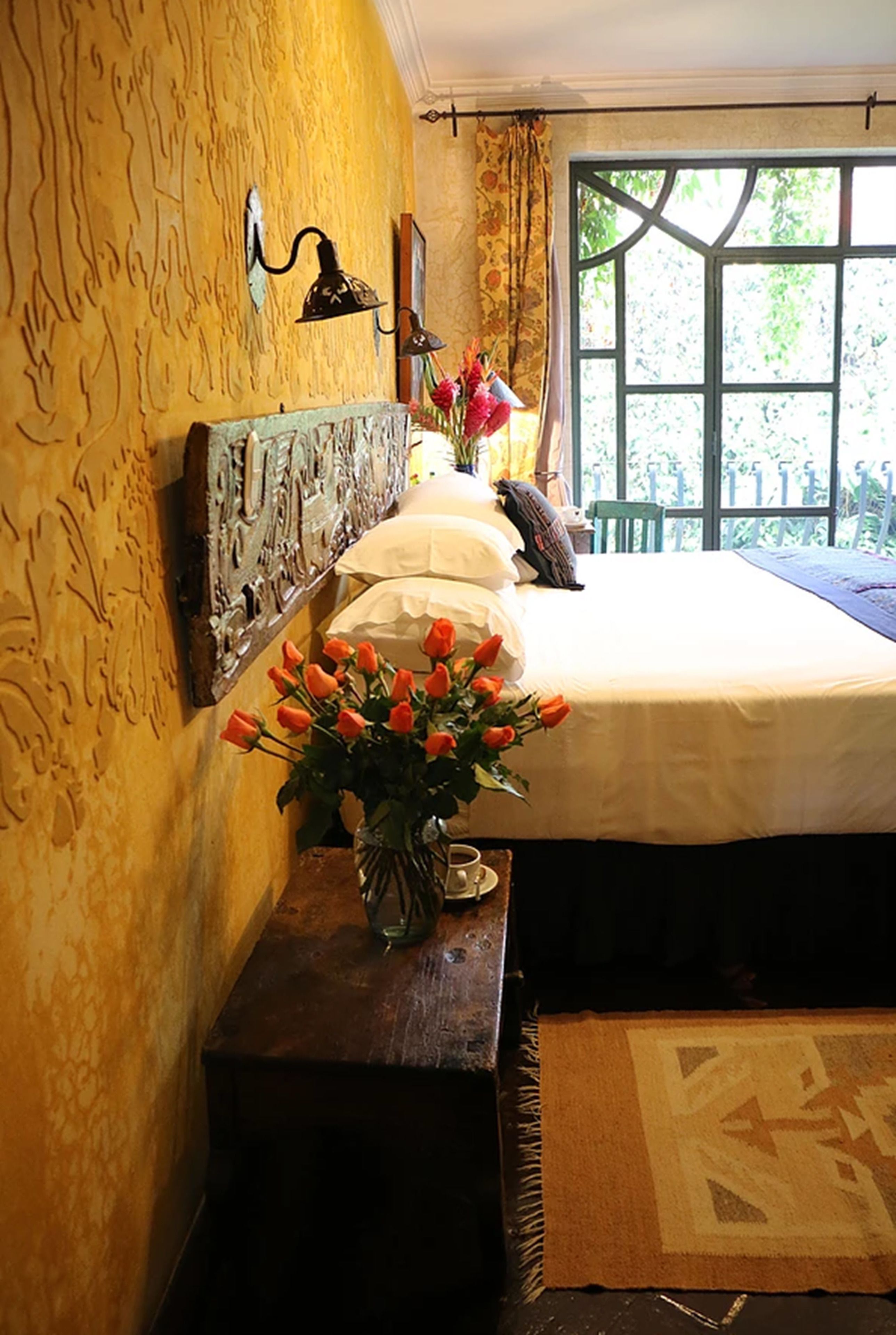 Une chambre à coucher avec un lit, une table de chevet ornée d’un vase de fleurs et une fenêtre avec des rideaux.