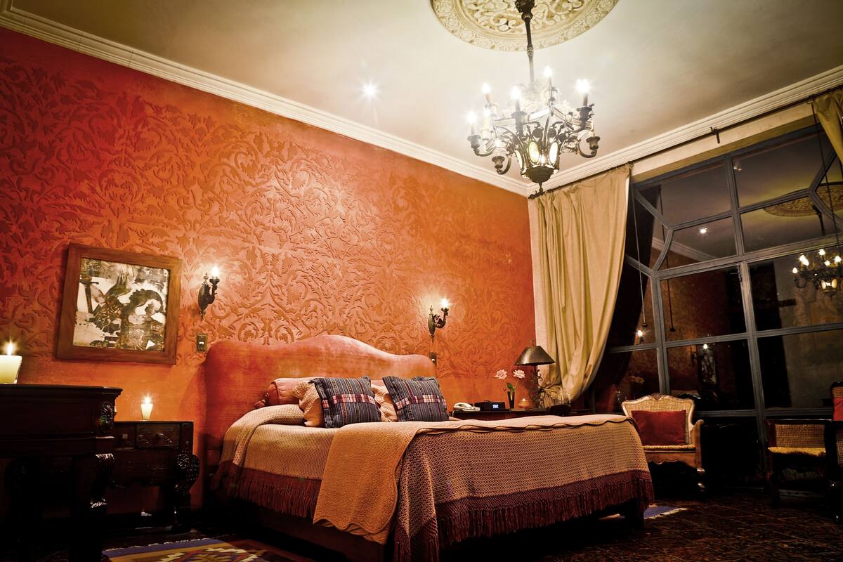 Une chambre à coucher avec un grand lit, un papier peint décoratif, un lustre et une grande fenêtre avec des rideaux.