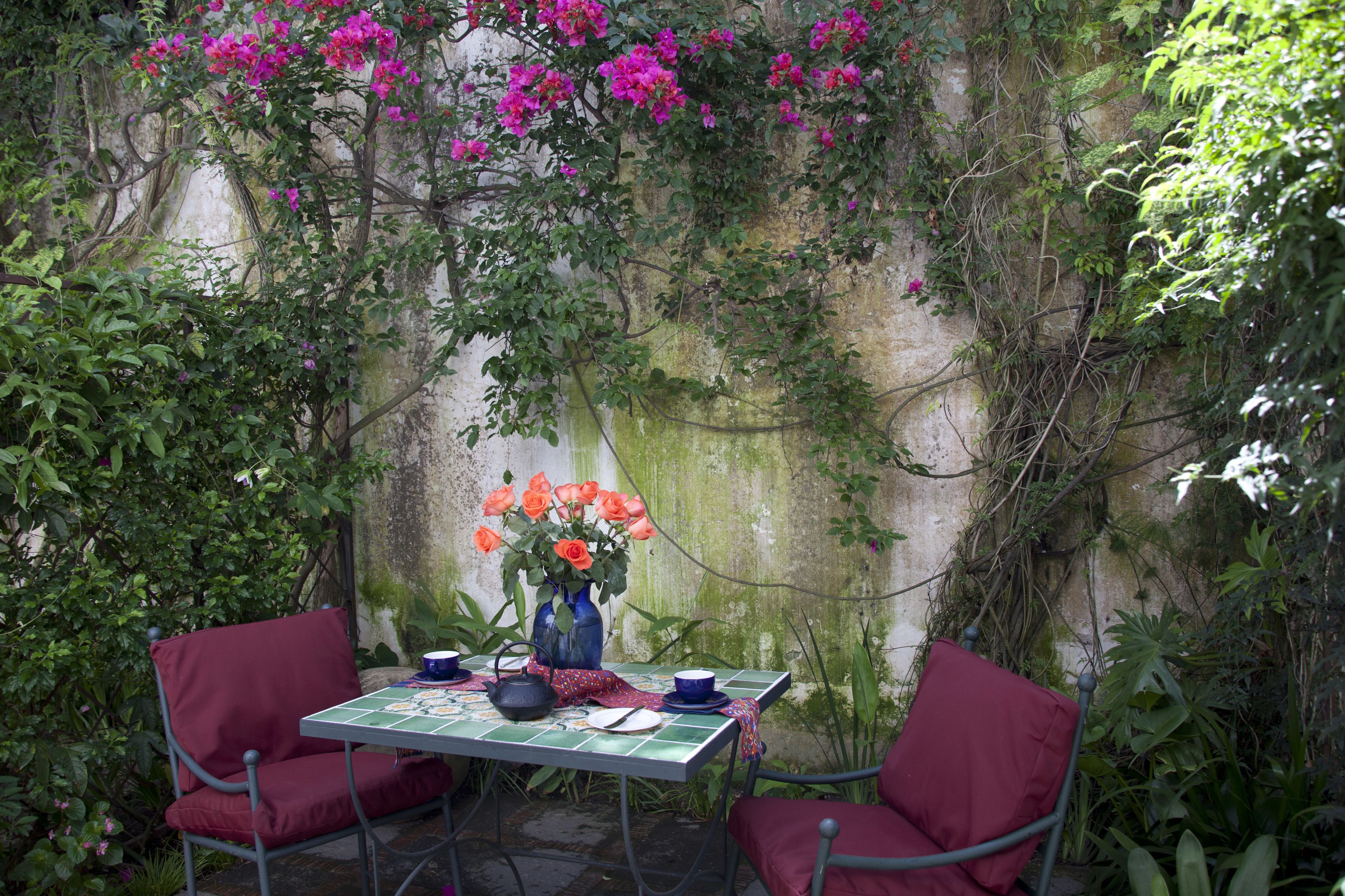 Ein Tisch mit Blumen, zwei Stühle und einer Tischdecke in einem Gartenbereich mit Steinmauer und viel Grün.