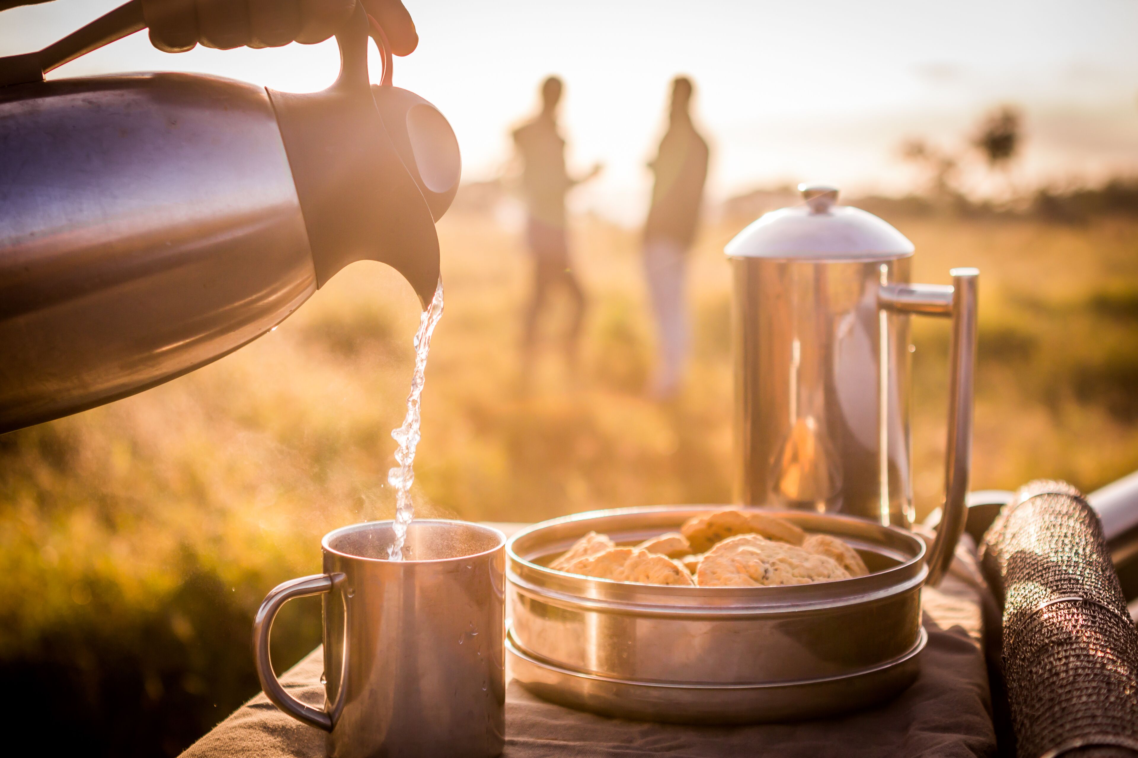 A stainless steel kettle pouring water into a mug, a thermos, and a metal container with food items on a table outdoors.