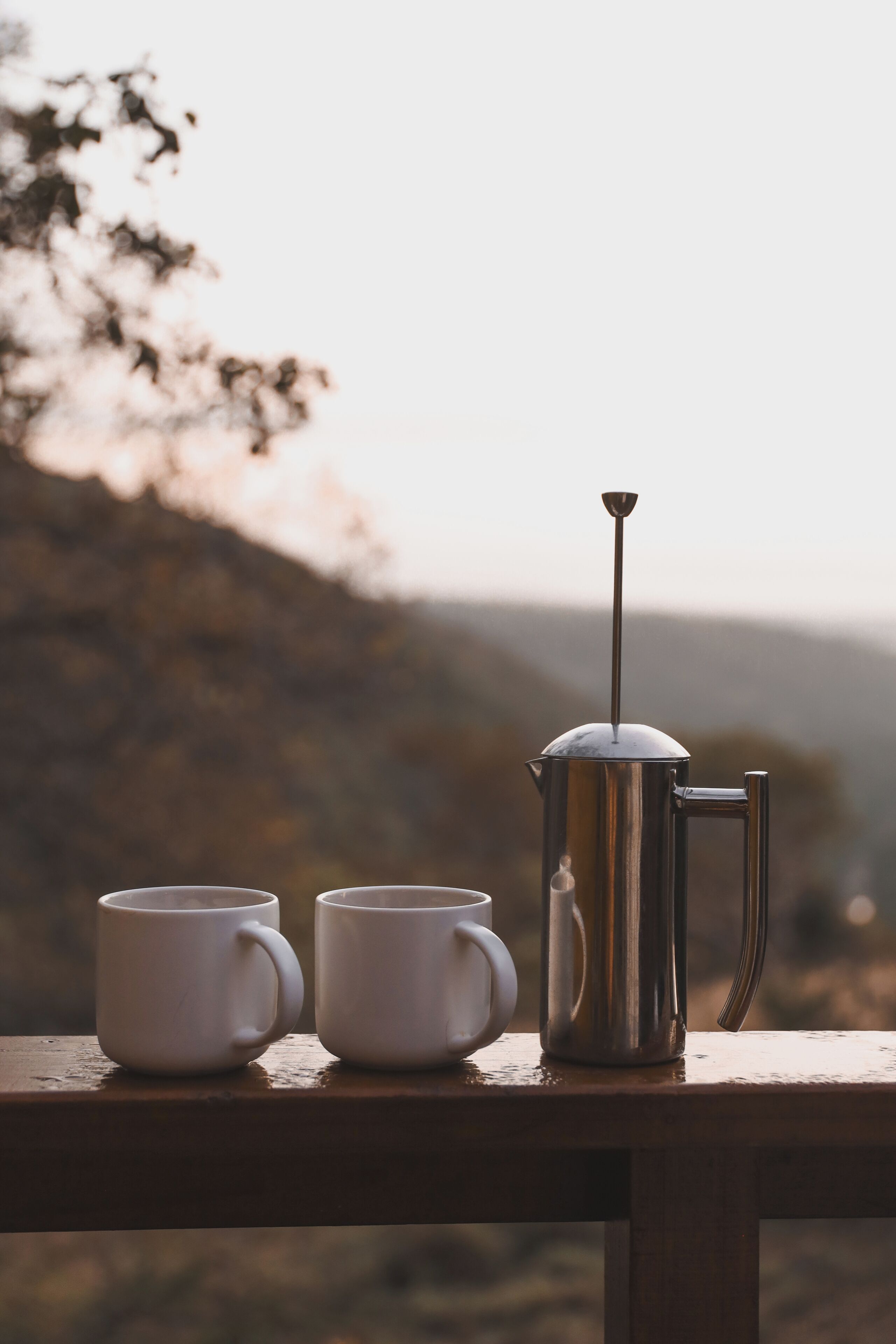 Uma cafeteira de metal e duas chávenas brancas sobre uma superfície de madeira, com um fundo ao ar livre desfocado.