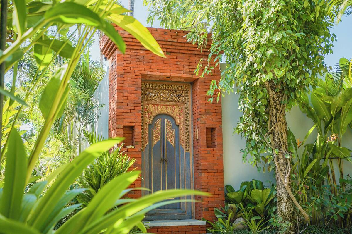 Un edificio de ladrillo tradicional con una puerta decorativa, rodeado de abundante vegetación.