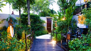 Un sentier en bois mène à un bâtiment doté d’une porte traditionnelle, le tout étant entouré d’une végétation luxuriante et de plantes ornementales.
