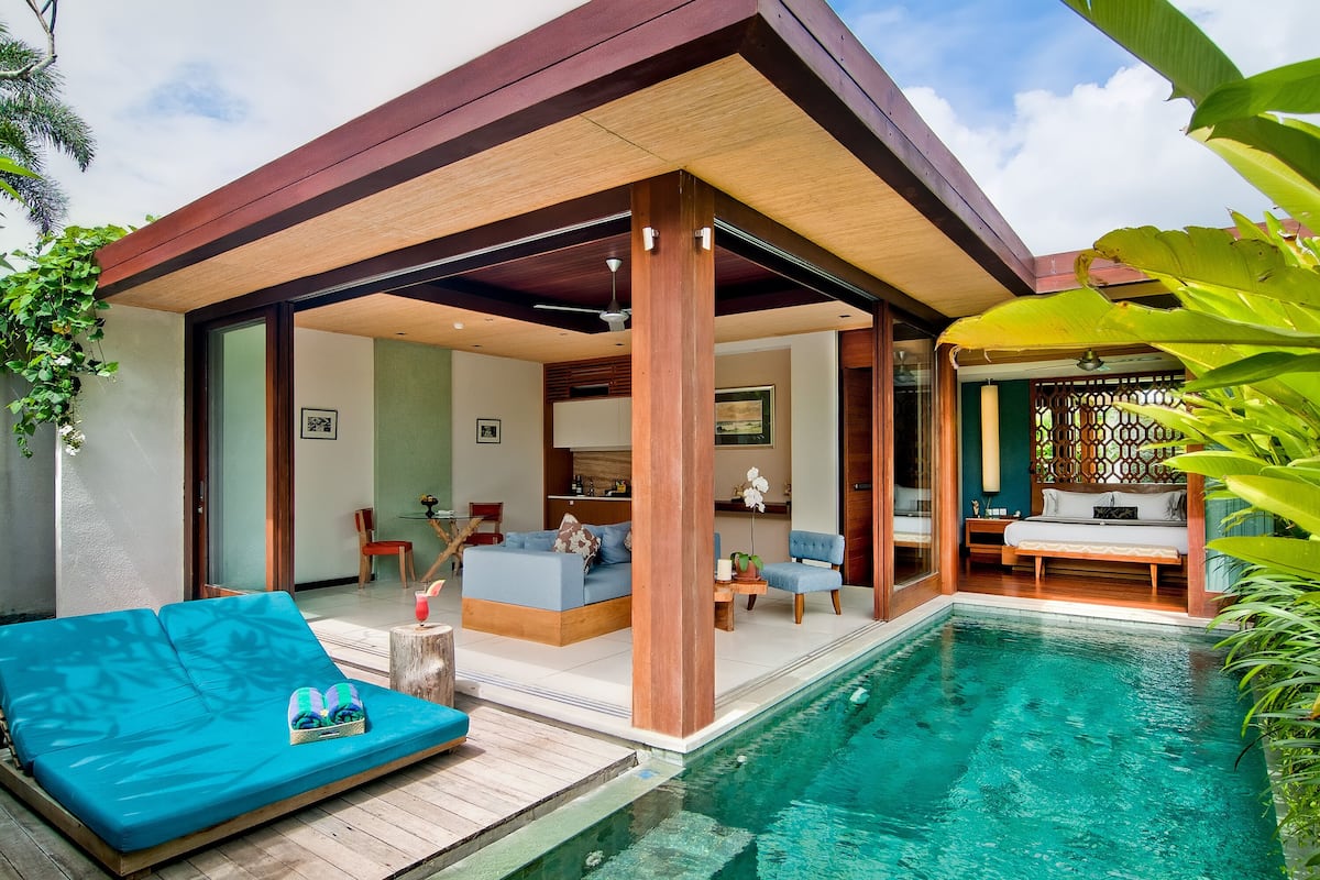Un área de piscina moderna al aire libre con una terraza de madera, un sofá azul y un banco de madera.