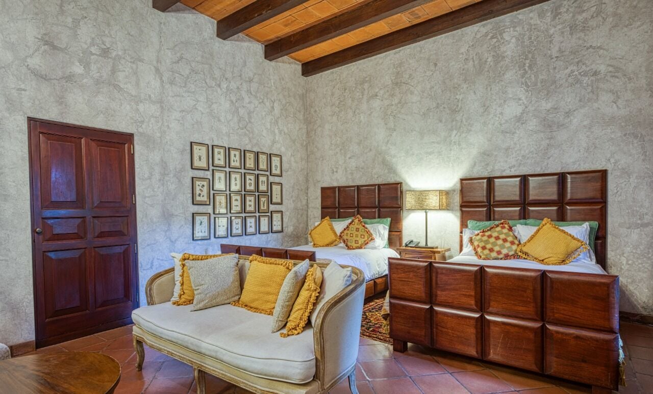 A bedroom with a large bed, a sofa, and a wooden cabinet. There are framed pictures on the wall and a wooden door.