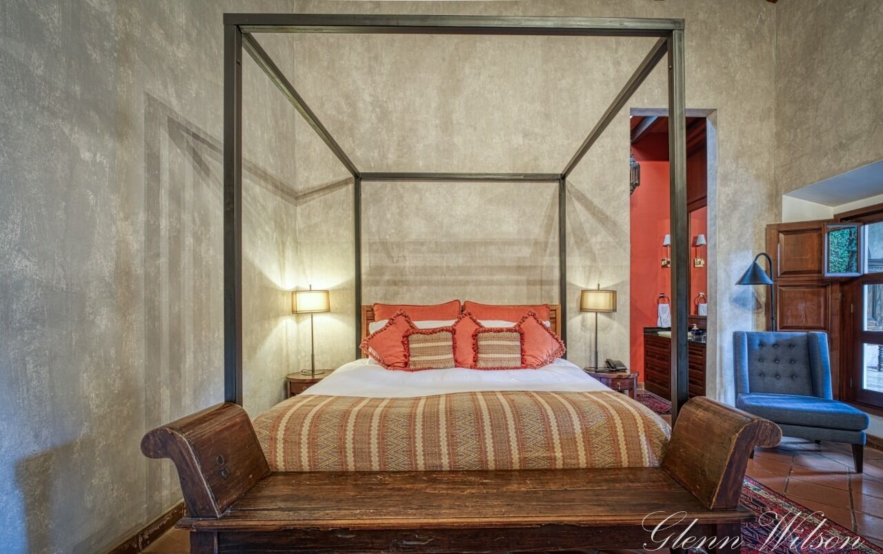 A four-poster bed with a wooden frame and a red and beige striped bedspread. There are two bedside tables with lamps, a blue upholstered chair, and a wooden bench at the foot of the bed.